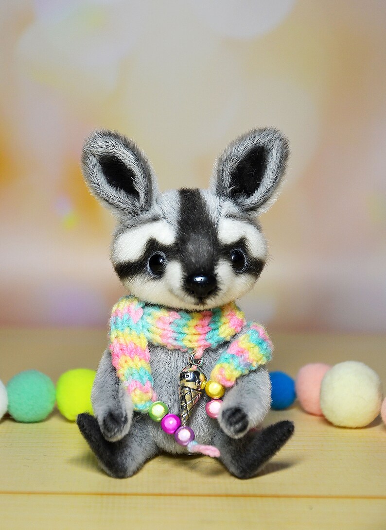 May include: A handmade plush raccoon with a gray and white face, black eyes, and large ears. It wears a colorful scarf and a charm necklace with an ice cream cone pendant. The raccoon is sitting with colorful pom-poms in the background.