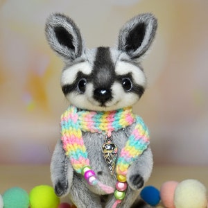 May include: A plush raccoon toy with a gray and white face, ears, and body. It wears a colorful rainbow scarf and a pendant with an ice cream cone charm. The toy has a black nose and big, dark eyes. A cute, handmade stuffed animal.