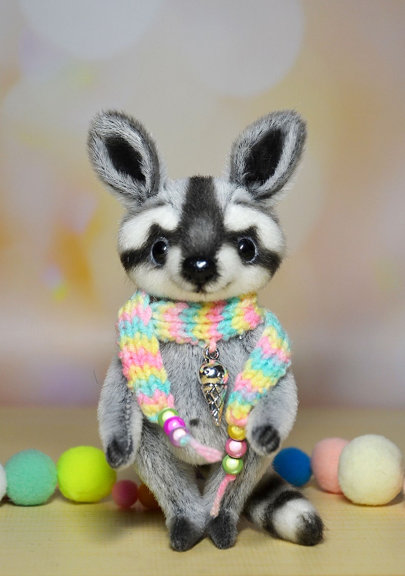 May include: A plush raccoon toy with a gray and white face, ears, and body. It wears a colorful rainbow scarf and a pendant with an ice cream cone charm. The toy has a black nose and big, dark eyes. A cute, handmade stuffed animal.