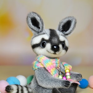May include: A plush raccoon doll with a gray and white face, ears, and striped tail. It wears a colorful scarf and a beaded necklace. The doll is surrounded by pastel-colored balls, creating a whimsical scene.