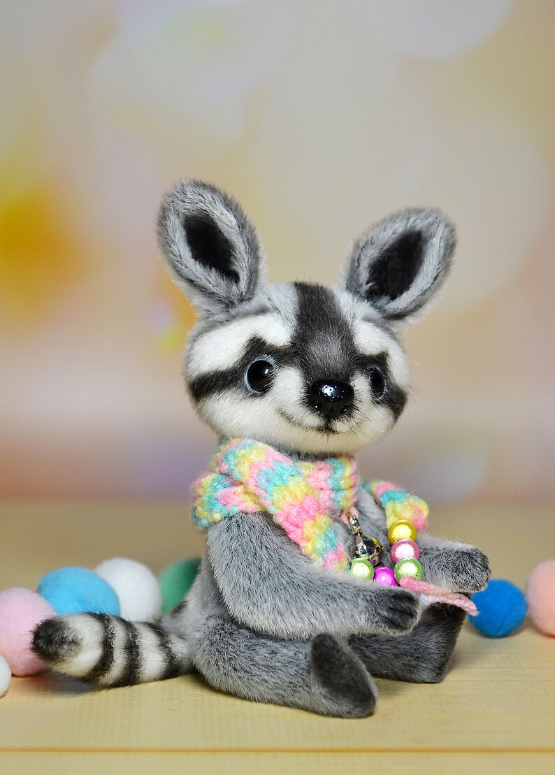May include: A plush raccoon doll with a gray and white face, ears, and striped tail. It wears a colorful scarf and a beaded necklace. The doll is surrounded by pastel-colored balls, creating a whimsical scene.