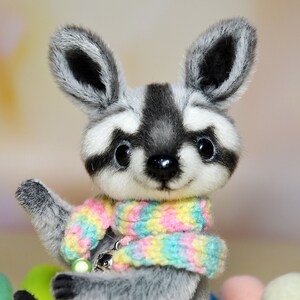 May include: A plush raccoon doll with a gray and white striped tail and a colorful scarf. The doll has large, expressive eyes and is holding its paw up. The scarf is pastel-colored, and the doll has a small charm attached.