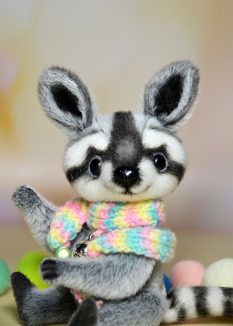 May include: A plush raccoon doll with a gray and white striped tail and a colorful scarf. The doll has large, expressive eyes and is holding its paw up. The scarf is pastel-colored, and the doll has a small charm attached.