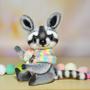 May include: A plush raccoon figurine with a gray and white striped tail and a colorful scarf. The raccoon has large, expressive eyes and is surrounded by colorful pom-poms. The figurine is designed to look like a stuffed animal.