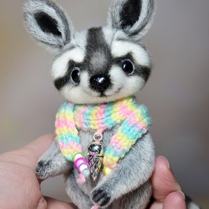 May include: A handmade plush raccoon with a black and gray striped tail and a colorful scarf. The raccoon has a silver ice cream cone charm around its neck. The plush animal is gray and white with large, dark eyes.