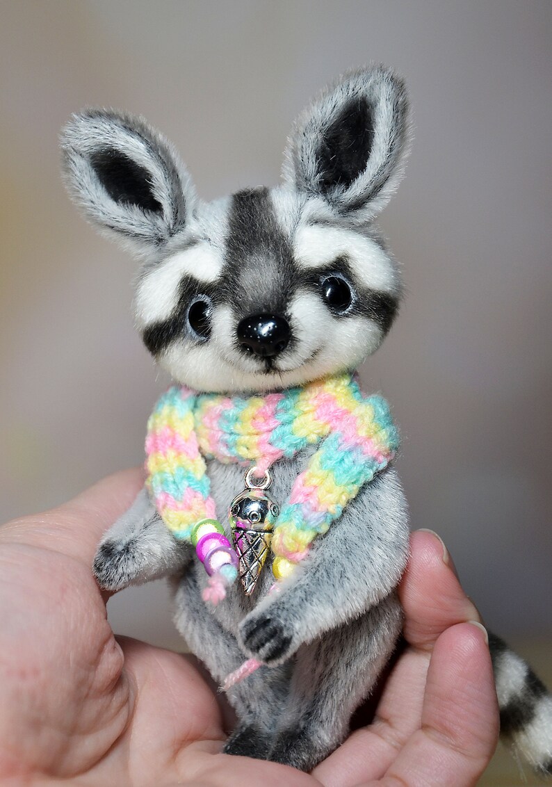 May include: A handmade plush raccoon with a black and gray striped tail and a colorful scarf. The raccoon has a silver ice cream cone charm around its neck. The plush animal is gray and white with large, dark eyes.