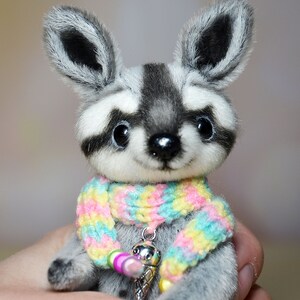 May include: A plush raccoon figurine with a gray and white face, large black eyes, and a colorful scarf. The raccoon has a small silver charm with an ice cream cone design. The figurine is a cute, handmade collectible.