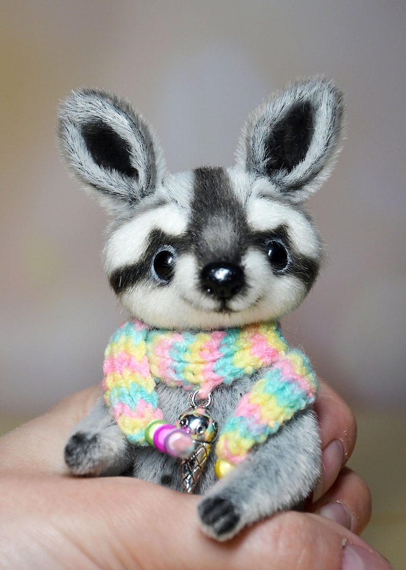 May include: A plush raccoon figurine with a gray and white face, large black eyes, and a colorful scarf. The raccoon has a small silver charm with an ice cream cone design. The figurine is a cute, handmade collectible.