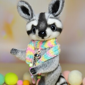 May include: A plush raccoon figurine with a black and white striped tail and a matching mask. It wears a colorful scarf and holds a charm of an ice cream cone. The figurine is surrounded by colorful pom-poms.
