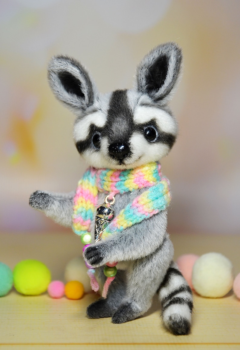 May include: A plush raccoon figurine with a black and white striped tail and a matching mask. It wears a colorful scarf and holds a charm of an ice cream cone. The figurine is surrounded by colorful pom-poms.