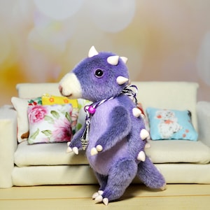 May include: A purple plush toy dinosaur with white horns and a white face. The dinosaur is wearing a pink and silver necklace and is standing on a white sofa.
