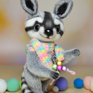 May include: A plush raccoon figurine with a gray and white striped body, wearing a colorful scarf and holding a beaded charm. The raccoon has large, dark eyes and a friendly expression. Decorative pom-poms in various colors are scattered around the base.