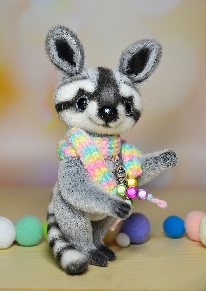 May include: A plush raccoon figurine with a gray and white striped body, wearing a colorful scarf and holding a beaded charm. The raccoon has large, dark eyes and a friendly expression. Decorative pom-poms in various colors are scattered around the base.
