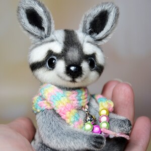 May include: A handmade plush raccoon with a gray and white striped face and ears. The raccoon wears a colorful scarf and a beaded necklace. The plush toy is held in a person's hand.