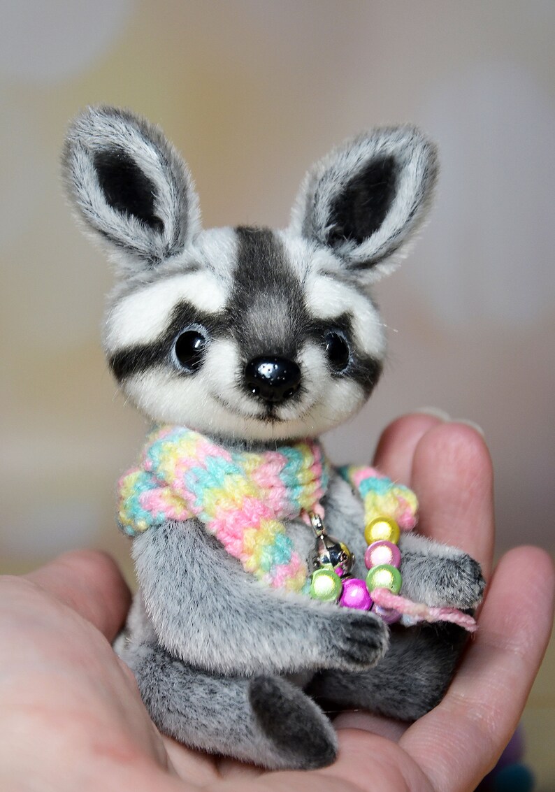 May include: A handmade plush raccoon with a gray and white striped face and ears. The raccoon wears a colorful scarf and a beaded necklace. The plush toy is held in a person's hand.
