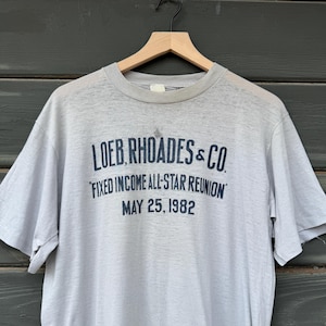 May include: A light gray vintage t-shirt with the text "LOEB RHOADES & CO." in dark blue. Below, it reads "FIXED INCOME ALL-STAR REUNION" and "MAY 25, 1982." The shirt is hanging on a wooden hanger.