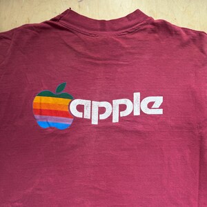 May include: A maroon-colored t-shirt featuring the Apple logo, a colorful apple design with a green leaf and a rainbow stripe pattern. The word "apple" is printed in white, block letters.