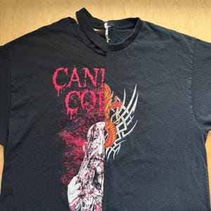May include: Black t-shirt with a split design. The left side features the text "CANI COR" in red dripping letters over a graphic of a face with red splatter. The right side has a tribal design.