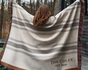 May include: Cream-colored woven blanket with horizontal black and gray stripes. The blanket has a brown border and colorful fringe. The text "THE COLES EST. 2024" is printed on the blanket.