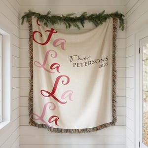 May include: A cream-colored throw blanket with the words "Fa La La La La" in a gradient of red and pink. The text "The Petersons 2025" is printed in black. The blanket has a fringed edge and is decorated with evergreen branches.