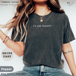 May include: A charcoal grey short-sleeve t-shirt featuring the white text I'll ask ChatGPT printed on the center chest. The garment has a crew neckline and a relaxed fit, paired with blue denim jeans and a gold-toned toggle pendant necklace.