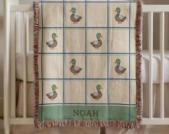 May include: A cream-colored baby blanket with a grid pattern and duck illustrations. The blanket has blue grid lines and a green border with the name "NOAH" in brown letters. The blanket is fringed on the edges.