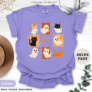 Pode incluir: Camiseta unissex violeta com um design tem&aacute;tico de Halloween. O design inclui gatos e fantasmas de desenhos animados em molduras laranja, com uma lua crescente e estrelas. A camiseta tem o texto "More Colors Available".