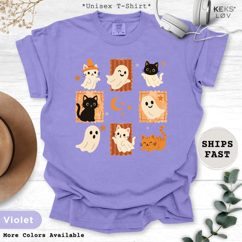 Pode incluir: Camiseta unissex violeta com um design tem&aacute;tico de Halloween. O design inclui gatos e fantasmas de desenhos animados em molduras laranja, com uma lua crescente e estrelas. A camiseta tem o texto "More Colors Available".