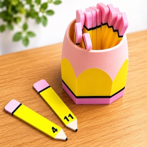 Op de afbeelding: Een potloodvormige bureau-organizer in roze en geel met een zwarte band, gevuld met diverse gele potloodvormige voorwerpen met roze gummen. Twee extra platte potloodvormige markers liggen ervoor, gemarkeerd met de cijfers 4 en 11.