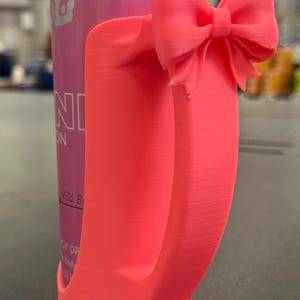 May include: A bright pink 3D-printed can holder with a bow detail. The holder is designed to fit a standard-sized can, and the bow adds a decorative touch. The can in the holder is pink with the text "Red Bull" and "Wild Berry".