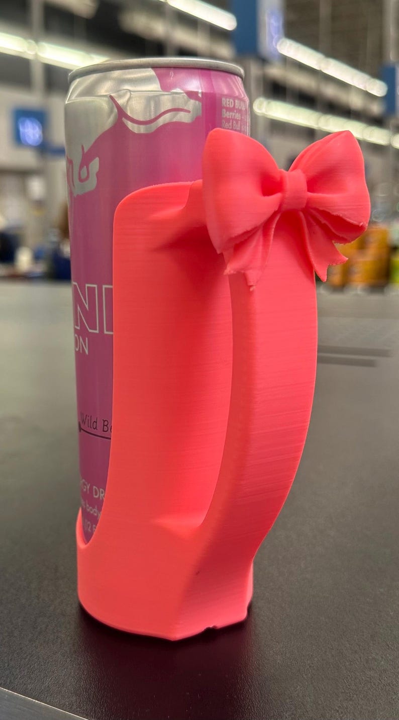 May include: A bright pink 3D-printed can holder with a bow detail. The holder is designed to fit a standard-sized can, and the bow adds a decorative touch. The can in the holder is pink with the text "Red Bull" and "Wild Berry".