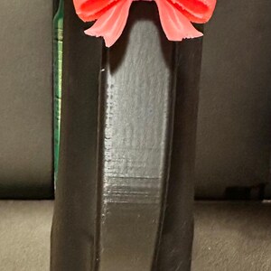 May include: A black, geometric-shaped object with a bright coral-colored bow attached to the top. The bow is detailed with multiple loops and a textured surface. The object appears to be 3D-printed.