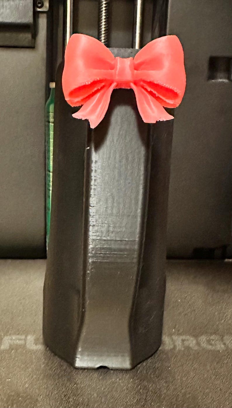 May include: A black, geometric-shaped object with a bright coral-colored bow attached to the top. The bow is detailed with multiple loops and a textured surface. The object appears to be 3D-printed.