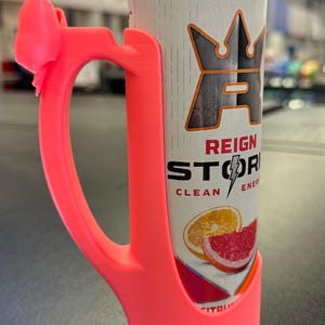 May include: A bright pink 3D-printed can holder with a bow-shaped accent on the handle. The holder encases a Reign Storm energy drink can, featuring citrus fruit graphics and the text "REIGN STORM CLEAN ENERGY".