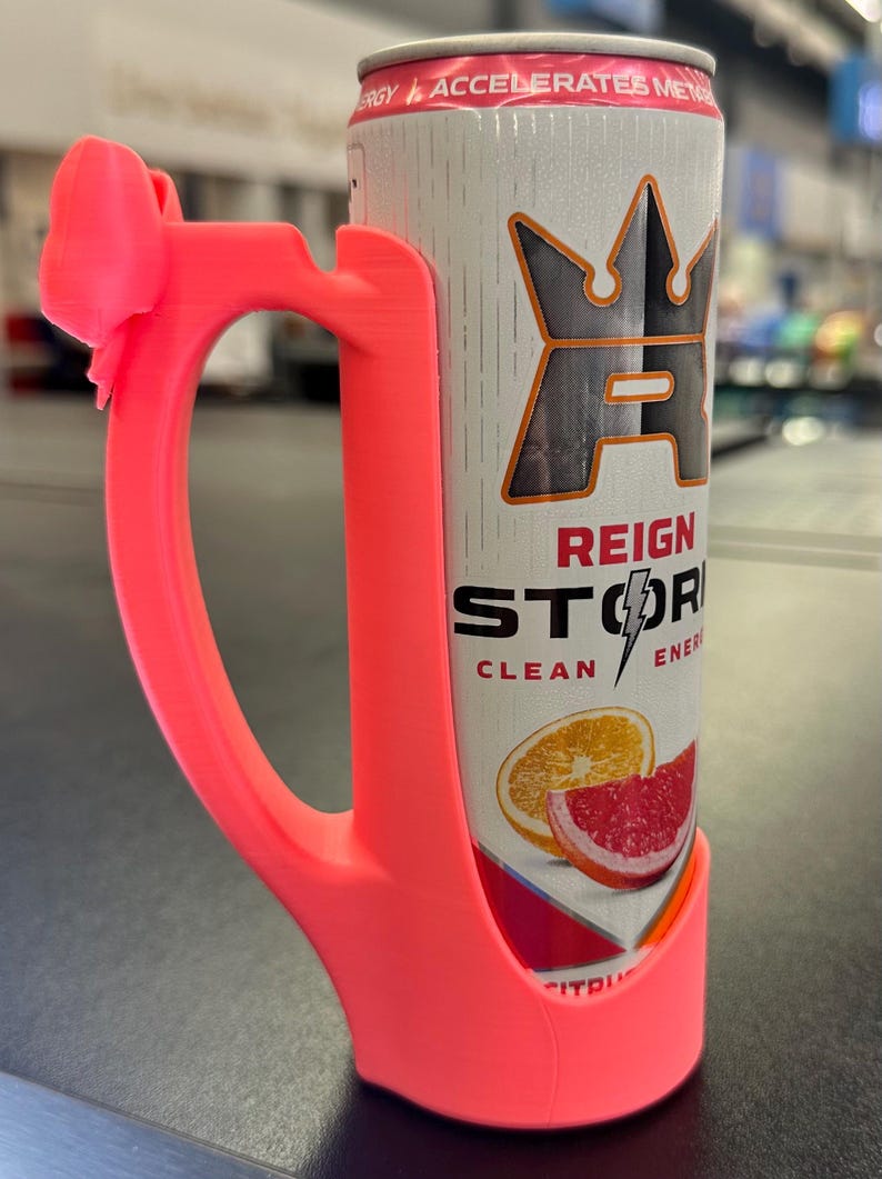 May include: A bright pink 3D-printed can holder with a bow-shaped accent on the handle. The holder encases a Reign Storm energy drink can, featuring citrus fruit graphics and the text "REIGN STORM CLEAN ENERGY".