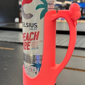 May include: A can of Celsius Peach Vibe sparkling white peach edition energy drink is held by a bright pink handle. The can has a white background with the Celsius logo and a palm tree design.