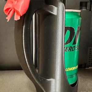 May include: A black 3D-printed can holder with a handle and a decorative pink bow attached. A green can of Zero Sugar Lemon-Lime soda is inside the holder. The holder is designed to keep beverages cold.