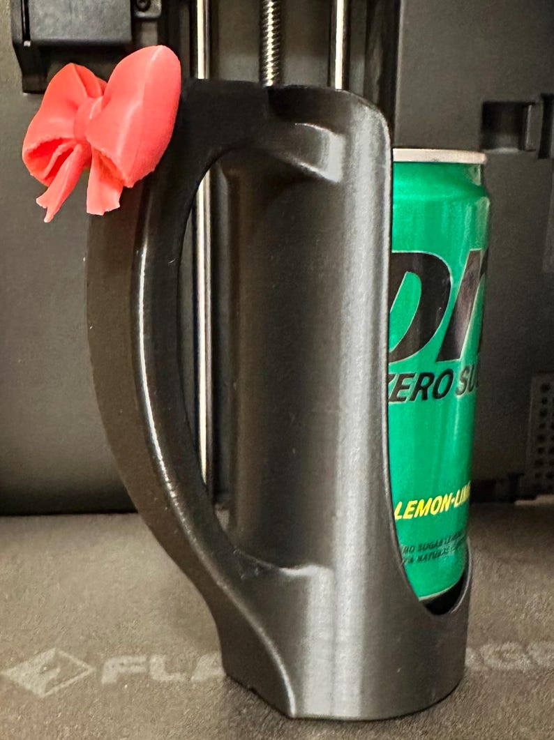 May include: A black 3D-printed can holder with a handle and a decorative pink bow attached. A green can of Zero Sugar Lemon-Lime soda is inside the holder. The holder is designed to keep beverages cold.