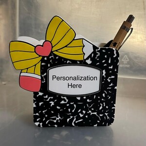 May include: A black and white composition notebook-style pen holder with a yellow pencil-shaped bow and a red heart. The holder has a white frame with the text "Personalization Here". A pen is inside the holder.