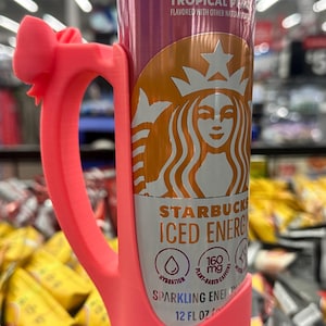 May include: A pink 3D-printed handle wraps around a Starbucks Iced Energy drink can. The can is primarily orange and purple with the Starbucks logo and the words "Tropical P" and "Sparkling Energy". The can is 12 fl oz.