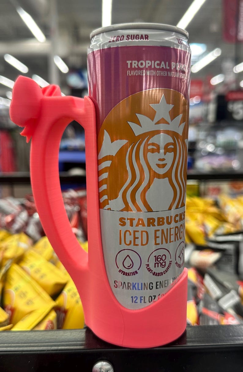 May include: A pink 3D-printed handle wraps around a Starbucks Iced Energy drink can. The can is primarily orange and purple with the Starbucks logo and the words "Tropical P" and "Sparkling Energy". The can is 12 fl oz.