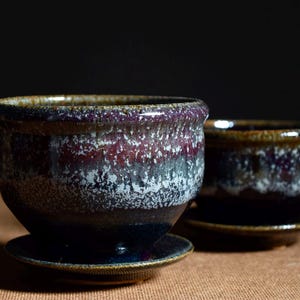 May include: Two ceramic bowls with matching saucers, featuring a reactive glaze that transitions from a deep dark blue base to a mottled white and vibrant purple midsection, finished with a smooth, rounded rim in a warm brown tone.