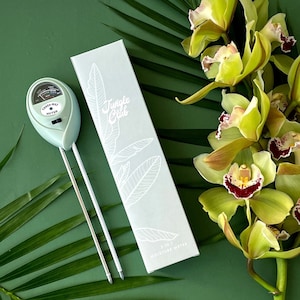 May include: A green and white 3-in-1 moisture meter with a dial and two probes. The meter is labeled "Three-Way Meter" and is packaged in a white box with the text "Jungle Club" and "3 in 1 Moisture Meter". The box is sitting on a green surface with green orchids.