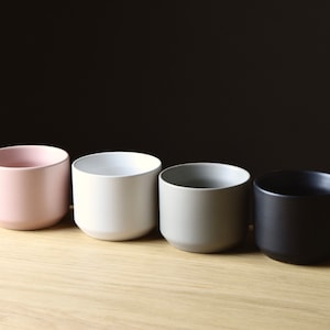 May include: Four ceramic planters in different colors: pink, white, gray, and black. The planters are cylindrical and have a smooth, matte finish.