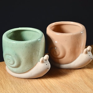 May include: Two ceramic snail planters, one green and one brown, with a speckled finish. The planters are shaped like snails with a small opening at the top for planting.
