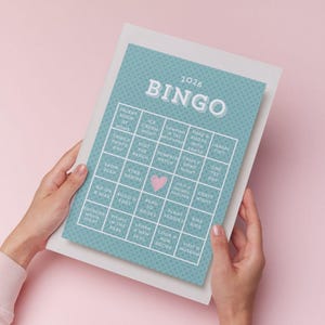 Puede incluir: Una tarjeta de Bingo turquesa de 2026 con un borde blanco es sostenida por manos sobre un fondo rosa. La tarjeta de Bingo tiene una cuadrícula de actividades como "Show de talentos en casa" y "Visitar la playa". La palabra "Bingo" está en la parte superior.