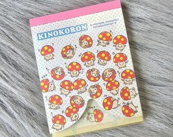 Puede incluir: Un bloc de notas rosa y amarillo con un diseño de hongo de dibujos animados. El bloc de notas tiene el texto "Kinokoron" y "*minnade tanoshiku korakorokoron*" en la parte delantera.
