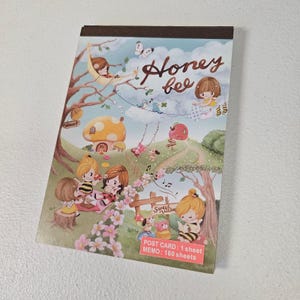 May include: A notepad with a whimsical illustration featuring cartoon characters, a mushroom house, and the words "Honey bee." The notepad has a brown top edge and includes the text "POST CARD: 1 sheet MEMO: 160 sheets."