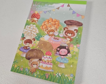 Puede incluir: Bloc de notas con una ilustración caprichosa que presenta personajes con forma de oso con disfraces de setas. La escena incluye una fiesta de té con pasteles y decoraciones coloridas, ambientada en un campo de hierba. El bloc de notas tiene el texto "kinoko no mori".