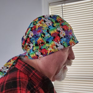 Könnte beinhalten: Eine bunte OP-Haube mit einem Muster aus Comic-Katzen. Die Haube ist am Hinterkopf gebunden und hat eine lange Schleife, die am Rücken herunterhängt.